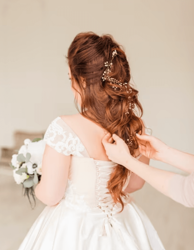 Foto di una sposa girata di spalle mentre la sua parrucchiera ritocca la coda morbida decorata da un motivo floreale 