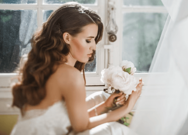 Una sposa fotografata con abito indossato, pettinatura sciolta e bouquet di fiori bianchi in mano che viene illuminata dalla luce della finestra 